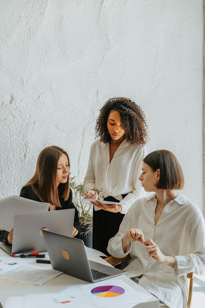 Three women collaborate on a project with charts and laptops in an office.